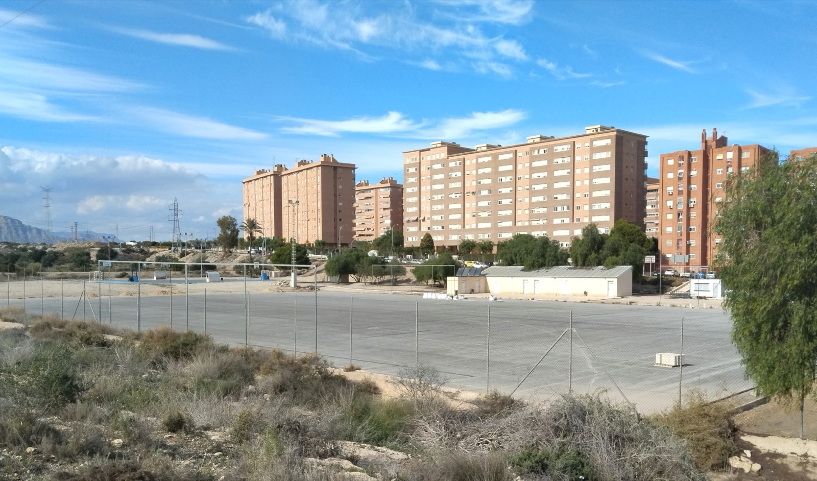 Campo municipal sin césped, con la base gris a la vista, una valla metálica y viviendas al fondo