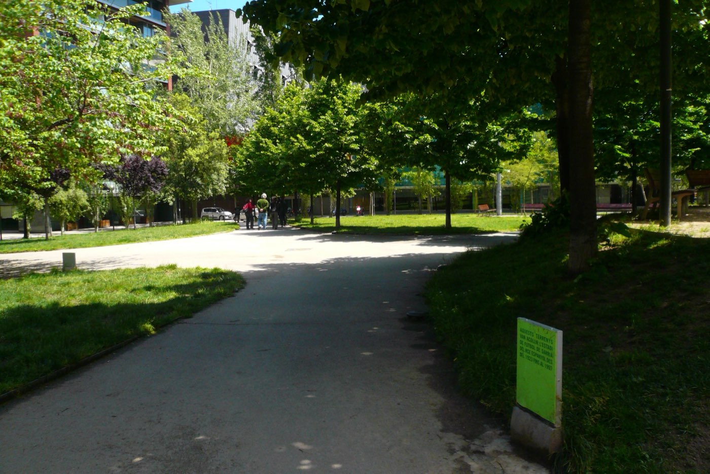 Camino entre césped y árboles en los jardines de Sarrià, con un cartel verde que recuerda el estadio